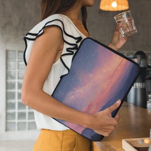 Puede incluir: Una funda negra para portátil con un diseño abstracto colorido de una puesta de sol o galaxia. La funda la sostiene una persona que lleva una blusa blanca con ribete negro y pantalones amarillos.