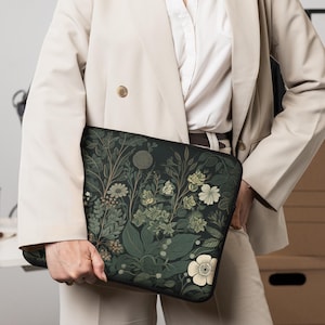 May include: A black laptop sleeve featuring a botanical design with white and green flowers, leaves, and stems. The floral pattern is set against a dark background. The sleeve is held by a person wearing a beige blazer.