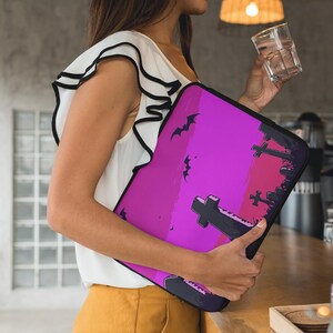 Puede incluir: Una funda para portátil con un diseño de Halloween. La funda tiene un fondo degradado de tonos rosa y morado, con siluetas negras de murciélagos y lápidas. La funda la sostiene una persona que lleva una camiseta blanca y pantalones amarillos.