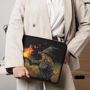Op de afbeelding: Zwarte laptoptas met een heks met een puntmuts, die een vuurbal vasthoudt. Het kunstwerk heeft een donkere achtergrond met gele en oranje accenten. De hoes wordt vastgehouden door een persoon die een beige blazer draagt.