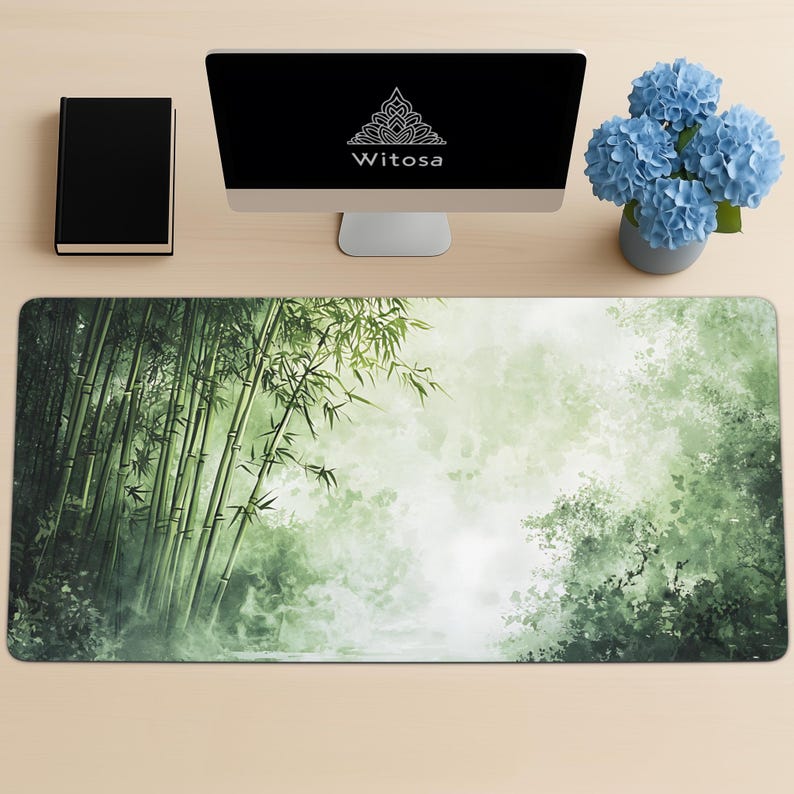 Desk mat with a green bamboo forest design, placed on a light wood desk with a computer, tablet, and blue hydrangea flowers in a gray pot.