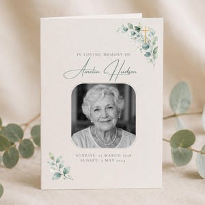 Puede incluir: Una tarjeta de programa funerario con un retrato en blanco y negro de una persona mayor, el texto IN LOVING MEMORY OF Amelia Hudson y las fechas SUNRISE: 12 MARCH 1938 SUNSET: 5 MAY 2024, decorada con hojas de eucalipto verdes en acuarela y una cruz dorada.