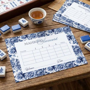 May include: A still-life shot of a Mahjong game setup. The scene includes a wooden tray with tiles, a small teacup, and scorecards. The scorecards have a blue floral design and the word "Mahjong Scorecard" printed on them.