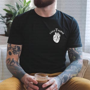 Op de afbeelding: Zwart t-shirt met een witte afbeelding van een hart en de tekst "CVICU NURSE". Het shirt heeft korte mouwen en een ronde hals. Het ontwerp is gecentreerd op de borst.