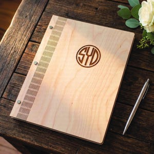 May include: A light-colored wooden binder featuring a vertical decorative laser-cut pattern along the left edge, secured with three silver metal fasteners. The front cover displays a circular monogram with the letters SYD engraved in the center.