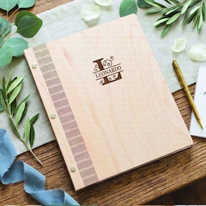 May include: A light-colored wooden binder with a vertical decorative strip featuring a repeating pattern and three metal fasteners. The cover is engraved with a large letter L and the name LEONARDO in a dark brown font centered on the front.