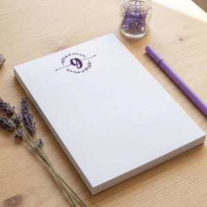 K&ouml;nnte beinhalten: Ein wei&szlig;er Notizblock mit einem violetten Buchstaben g, der in einem kreisf&ouml;rmigen botanischen Kranz zentriert ist. Der Notizblock wird von einem violetten Stift, einem kleinen Glas mit violetten B&uuml;roklammern, einem Amethystkristall und Lavendelzweigen begleitet.