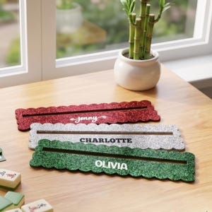 Può includere: Tre supporti espositivi rettangolari glitterati con bordi smerlati, ciascuno dotato di una fessura orizzontale. Il supporto rosso riporta il nome Jenny, quello argento Charlotte e quello verde il nome Olivia in caratteri bianchi.