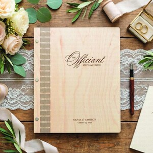 May include: A wooden wedding officiant book with the text "The Officiant" and the name "Stephanie Smith" engraved on the cover. The book has a decorative vertical stripe and is surrounded by flowers and wedding rings.