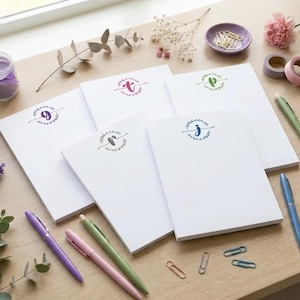 May include: Five white rectangular notepads, each featuring a unique lowercase letter monogram in a circular floral wreath design. The letters include g, t, p, r, and j in various colours. The notepads are arranged with pens, paper clips, and flowers.