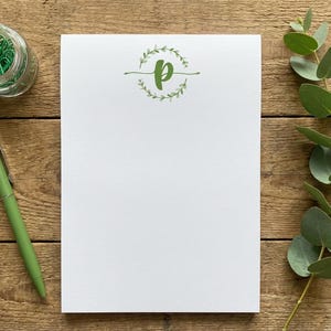 K&ouml;nnte beinhalten: Ein wei&szlig;er Notizblock mit einem gr&uuml;nen Buchstaben P, der in einem kreisf&ouml;rmigen botanischen Kranz zentriert ist. Der Notizblock wird von einem gr&uuml;nen Kugelschreiber und einem mit gr&uuml;nen B&uuml;roklammern gef&uuml;llten Glas begleitet, neben einem Eukalyptuszweig.