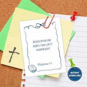 May include: A white card with the text "REJOICE IN THE LORD ALWAYS. I WILL SAY IT AGAIN: REJOICE!" and "Philippians 4:4" in blue. The card is held by a red paper clip and pinned to a corkboard with a red push pin.