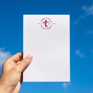K&ouml;nnte beinhalten: Eine rechteckige wei&szlig;e Schreibwarenkarte mit einem dunkelrosa Kleinbuchstaben t, der oben zentriert ist und von einem zarten kreisf&ouml;rmigen Blattkranz sowie feinen horizontalen Linien an den Seiten des Buchstabens umschlossen wird.