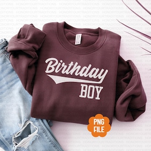 Puede incluir: Una sudadera burdeos con el texto "Birthday BOY" en blanco, con una fuente de estilo béisbol. La sudadera está doblada sobre un par de vaqueros azules. Un icono de archivo PNG está en la esquina inferior derecha.