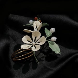 May include: A floral hair comb featuring cream-colored flower petals with a central crystal accent. The comb includes green leaves, white beaded accents, and a gold-toned metal comb base. The background is a dark, textured fabric.