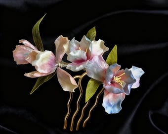 Op de afbeelding: Een bloemen haar kam met delicate, pastelkleurige bloemen in roze, blauw en wit, met groene bladeren. De bloemen zijn bevestigd aan een goudkleurige metalen kam, perfect voor bruiloften of speciale gelegenheden.