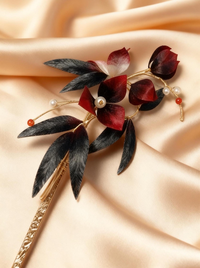 May include: A decorative hair accessory featuring a floral design. The piece has dark red petals, black leaves, and small white pearls. The stem and base are gold-colored, and the accessory is set against a satin background.