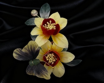Könnte beinhalten: Florale Brosche mit zwei gelb-roten Blüten, detaillierten Blütenblättern und zentralen Staubblättern. Die Brosche umfasst dunkelgraue und grüne Blätter, einen Schmetterling und einen funkelnden Akzent. Der Hintergrund ist schwarz.