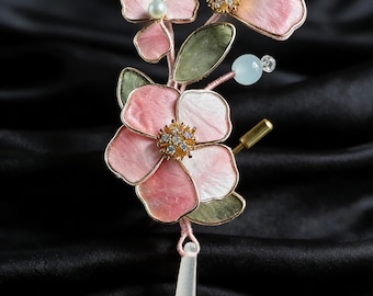Könnte beinhalten: Blumenbrosche mit rosa und weißen Blütenblättern mit goldfarbenen Rändern. Sie hat grüne Blätter, einen hängenden rosa Tropfen, kleine Kristalle und eine Perle. Auf schwarzem Hintergrund.