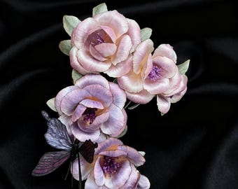 Könnte beinhalten: Eine handgefertigte Blumenbrosche mit vier zarten, hellvioletten und rosa Stoffblumen mit detaillierten Blütenblättern und violetten Zentren. Eine dunkelviolette und schwarze Libelle ist an den Blumen befestigt. Der Hintergrund ist schwarz.