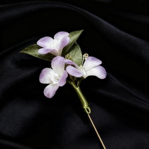 May include: A floral boutonniere with three pale purple and white flowers and green leaves. The stems are wrapped in green thread, with a gold pin extending from the base. Set against a black, silky fabric.