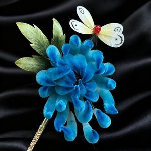 May include: A vibrant blue velvet flower with a gold stem and green leaves, alongside a white butterfly with a red centre. The flower has a detailed texture, set against a black backdrop.