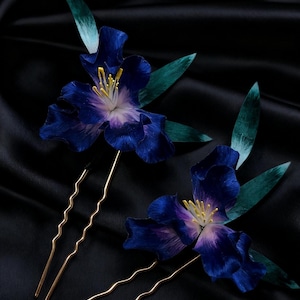 以下が含まれることがあります： 金色の茎が付いた、濃い青色の花のヘアピン2つ。 花びらは紫と青のグラデーションで、中央には黄色と白のディテールがあります。 緑の葉がコントラストを加えています。