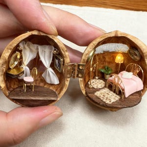 May include: A detailed miniature bedroom scene housed within a walnut shell. The interior includes a bed with a pink blanket, a small chair, a vanity, and a lamp. The walnut shell is open, showcasing the handcrafted details.