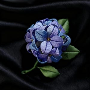 以下が含まれることがあります： 青と紫の色合いの手作りの刺繍アジサイの花、緑の葉と茎付き。 花は布と糸でできており、黒い背景に配置されています。