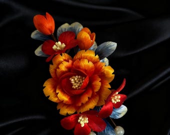 May include: A handcrafted floral hair accessory featuring a large, multi-layered orange and yellow flower with smaller red and orange blooms. Blue and white leaves and pearl accents complete the design, set against a black background.
