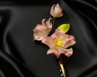 May include: A delicate floral boutonniere featuring three light pink flowers with yellow centers and a single green leaf. The flowers are attached to a brown stem and secured with a gold-colored clip. The background is a black satin fabric.