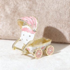 May include: A decorative baby carriage figurine in white, pink, and gold. The carriage features a pink canopy with crystal accents, pink wheels, and a gold handle. The carriage is a charming keepsake.
