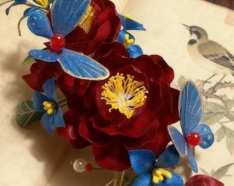 Op de afbeelding: Een handgemaakt bloemen haaraccessoire met dieprode fluwelen bloemen met gele centra, blauwe vlinderaccenten en kleine blauwe bloemen. Het accessoire is versierd met parels en rust op een open boek met een vogelillustratie.