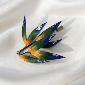 May include: A leaf-shaped brooch with a gold pin. The leaves are painted in blue, green, and orange, with gold leaf accents. The brooch is set against a white background.