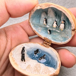 May include: A miniature diorama inside a hinged walnut shell featuring a polar scene. The top half shows three small penguins on an icy ledge with white peaks. The bottom half depicts a blue water pool with two swimming penguins and a snowy shore.