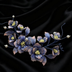Puede incluir: Un accesorio floral para el cabello con flores azules y rosas, centros amarillos y detalles de perlas. Las flores están dispuestas sobre una base de alambre azul oscuro, creando un diseño elegante y delicado. El fondo es de tela de satén negro.