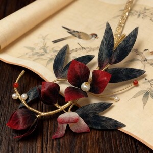 May include: A decorative hair accessory featuring a gold-toned metal frame with dark red and black leaves and flowers. The design includes faux pearls and red beads. The accessory rests on a scroll with a bird illustration.