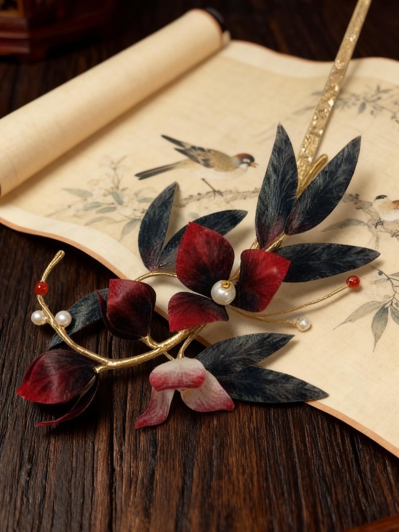 May include: A decorative hair accessory featuring a gold-toned metal frame with dark red and black leaves and flowers. The design includes faux pearls and red beads. The accessory rests on a scroll with a bird illustration.
