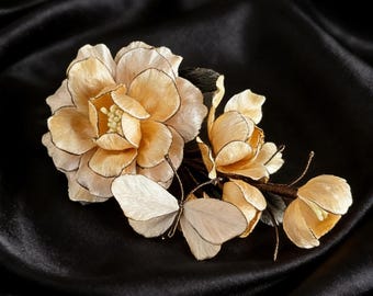 Könnte beinhalten: Handgefertigtes Haaraccessoire mit einer großen, hellpfirsichfarbenen Blüte mit schwarzen Rändern, begleitet von kleineren Blüten und Schmetterlingsakzenten. Auf dunklem, seidigem Stoff.