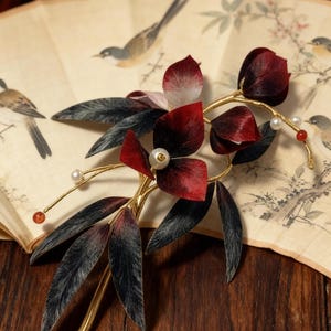May include: A decorative hair accessory featuring burgundy fabric flowers and dark gray leaves on a gold-colored stem. Small pearls and red beads accent the design. The accessory rests on a vintage fan with bird illustrations.