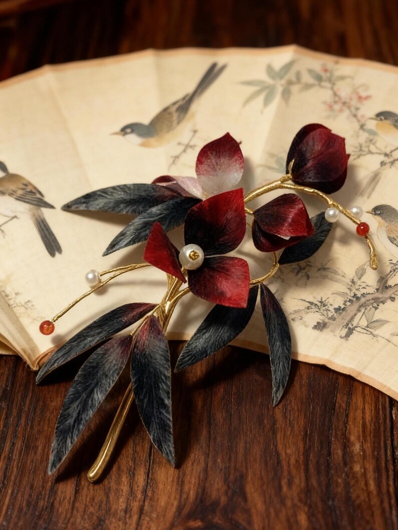 May include: A decorative hair accessory featuring burgundy fabric flowers and dark gray leaves on a gold-colored stem. Small pearls and red beads accent the design. The accessory rests on a vintage fan with bird illustrations.