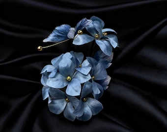 Könnte beinhalten: Ein zartes blaues Blumen-Haaraccessoire mit einer Ansammlung von Stoffblumen mit goldenen Akzenten. Ein blauer Schmetterling mit goldenen Details ist an den Blumen befestigt. Das Accessoire ist vor einem schwarzen Satinhintergrund platziert.