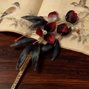 May include: A decorative hair accessory featuring a floral design with red and black petals, black leaves, and faux pearls. The gold-colored stem and base are intricately detailed. The background includes a vintage book with bird illustrations.