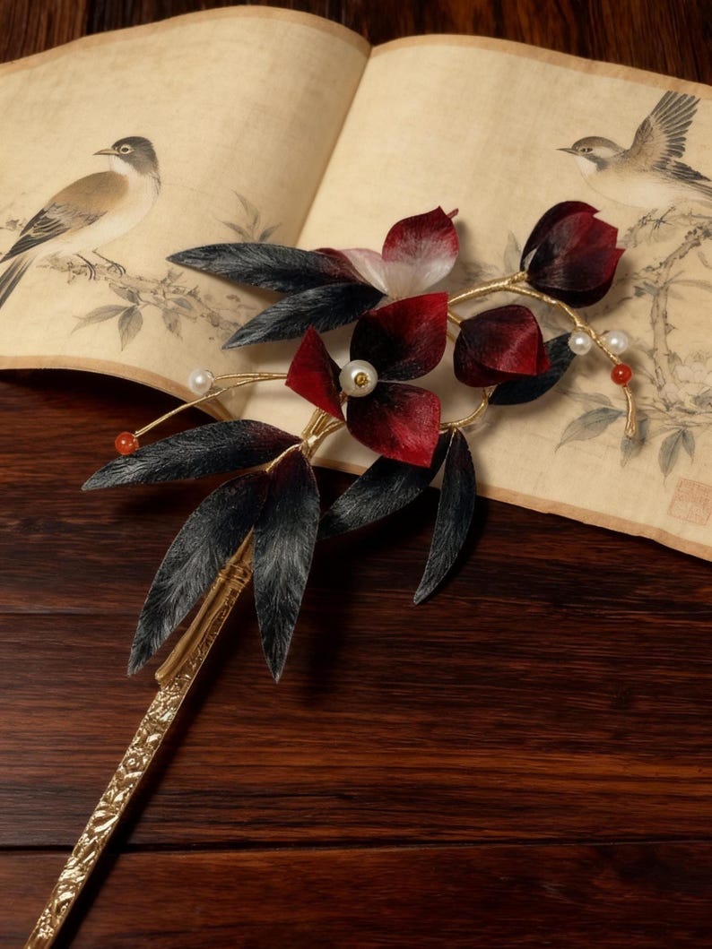 May include: A decorative hair accessory featuring a floral design with red and black petals, black leaves, and faux pearls. The gold-colored stem and base are intricately detailed. The background includes a vintage book with bird illustrations.
