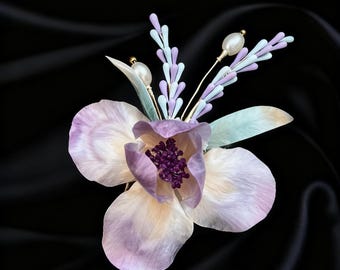 May include: A handcrafted floral brooch featuring a large, light purple flower with a dark purple center and delicate petals. Accented with pale blue and lavender sprigs, gold wire, and pearl-like beads. Set against a black background.