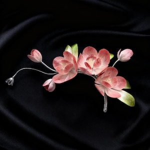 May include: A floral hair accessory with pink blossoms and green leaves. The flowers are attached to a silver-coloured wire, creating an elegant design. The accessory is set against a black background.