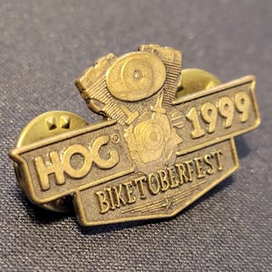 May include: A vintage brass-colored pin featuring a motorcycle engine design. The pin reads "HOG" and "BIKETOBERFEST 1999". The pin has a classic, aged appearance, suggesting it is a collectible item.
