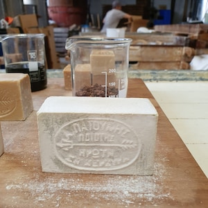 May include: A white bar of soap with the text "ΠΑΤΟΥΧΑΣ ΠΟΛΙΤΗΣ" and "ΠΡΩΤΗ ΚΕΡΚΥΡΑ" in Greek. The soap is on a wooden surface with white powder on it.