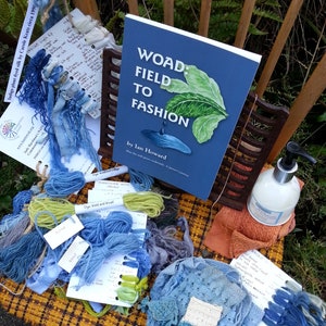 May include: A book titled "Woad: Field to Fashion" by Ian Howard, featuring a green leaf design on the cover, is shown on a wooden chair with assorted blue yarn, fabric scraps, and a bottle of liquid.
