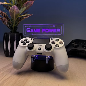 May include: A white PlayStation 4 controller is displayed on a black stand with a purple LED sign that reads "GAME POWER".